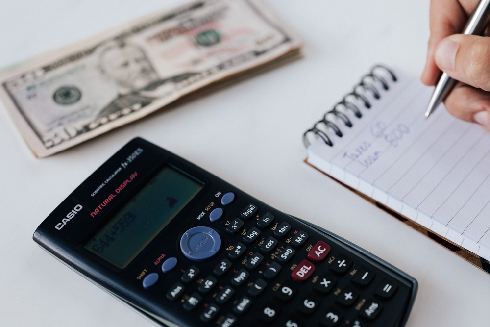 An image of a calculator, a person's hand writing in a notepad and stack of cash that represents creating a travel budget