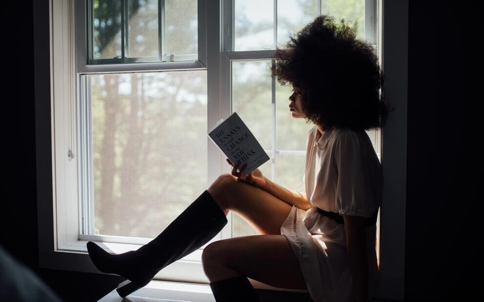 a woman sitting at a window reading a book. the featured image for a blog post about the most important areas to simplify in your life. 