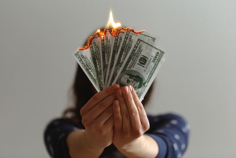 A woman holding up a stack of hundred dollar bills that are on fire. This image represents money saving tips to avoid