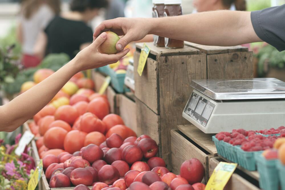 An image of two hands. One is handing a piece of fruit to the other. The background is of a farmer's market. This image represents food, which is one of the areas you should account for in a travel budget