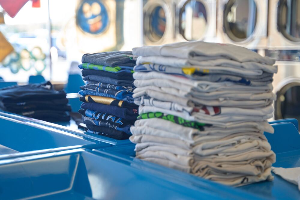 An image of a folded stacks of clothes at a laundry mat which represents using less detergent which is one of many unusual frugal tips shared in this blog post.