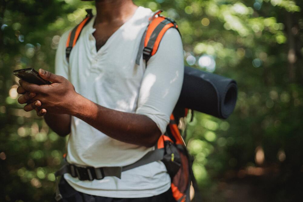 A man holding a phone in the wilderness. He has a backpacking backpack on. This image represents communication which is one of the areas you want to account for in your travel budget