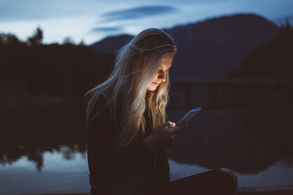 a girl scrolling on her phone by a lake. it is dark outside and only her phone is illuminated by light