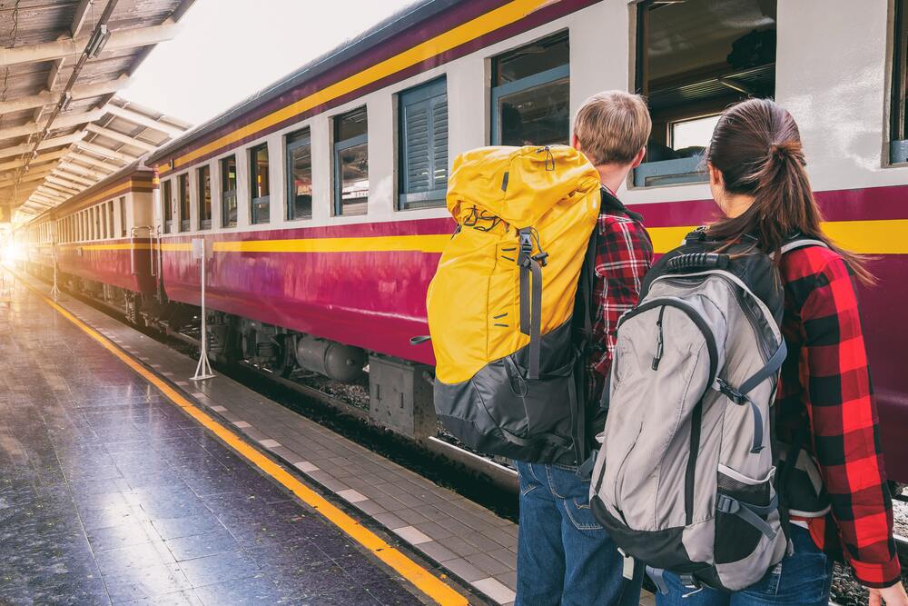 Two people wearing backpacks and waiting at the train station. This image represents transportation within the destination which is one of the areas you should account for in your travel budget