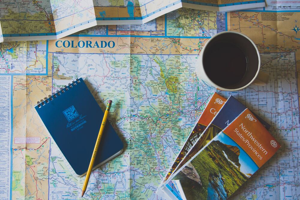 A map of Colorado with a coffee cup, notepad and brochures on top of it that represents the process of creating a travel budget