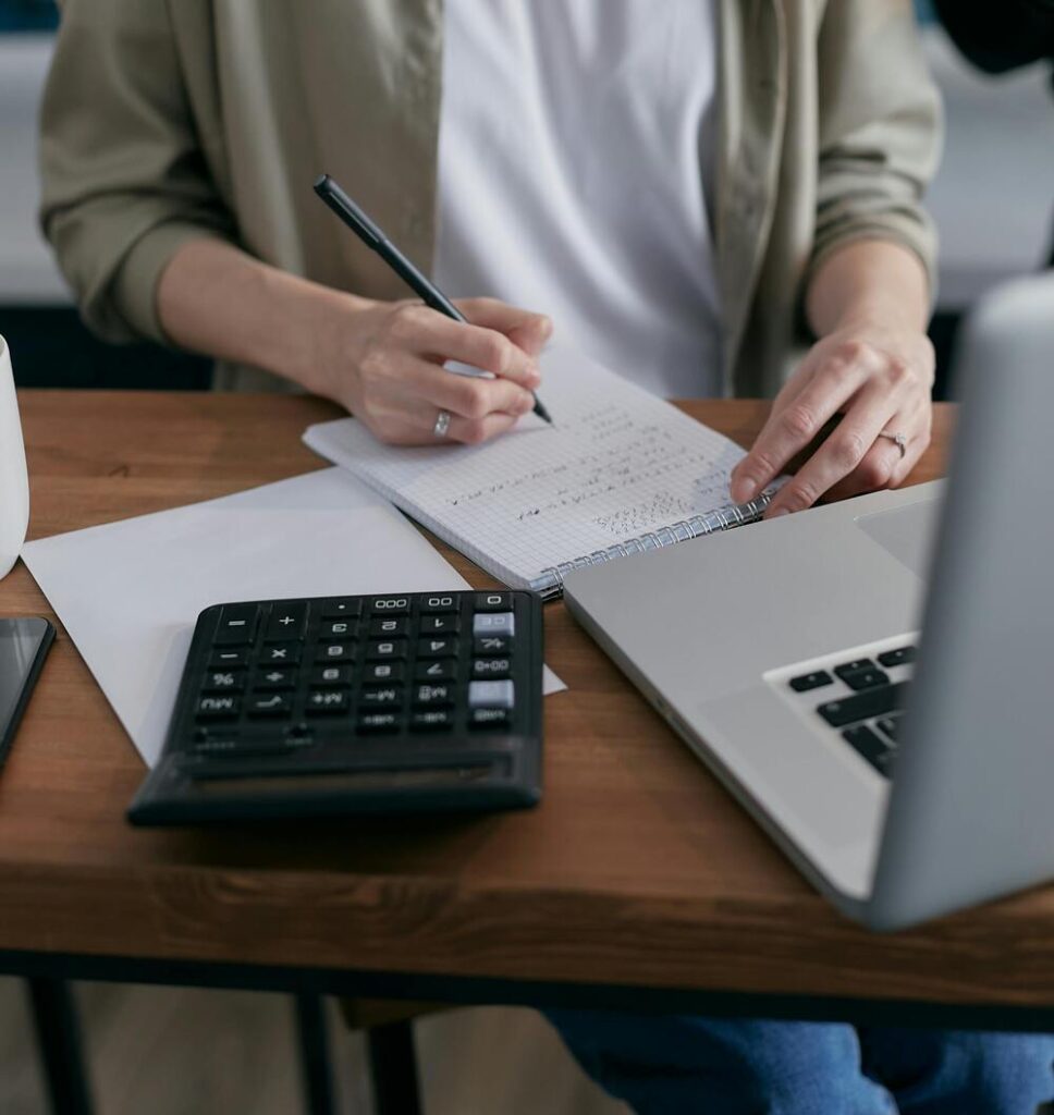 A woman in a gray blazer and white t-shirt writing in a notepad. There is a laptop and calculator on the wooden table that she is working at. This image represents budgeting methods