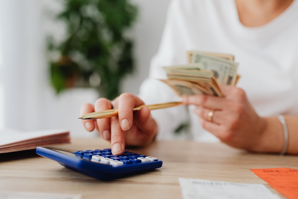 A close up image of a woman in a white shirt counting her cash. She has a wad of cash in one hand and a pen and calculator in the other. This image represents budgeting using budgeting methods. 