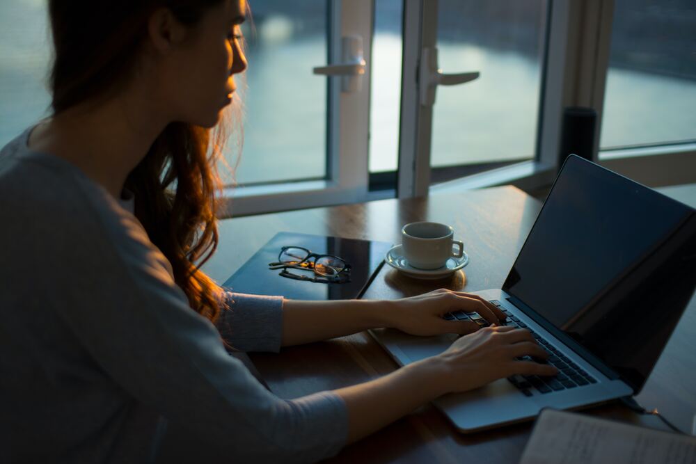A woman sitting at her computer looking at a blog about digital minimalism. There is a coffee cup, notebook and glasses on the table she works at. 