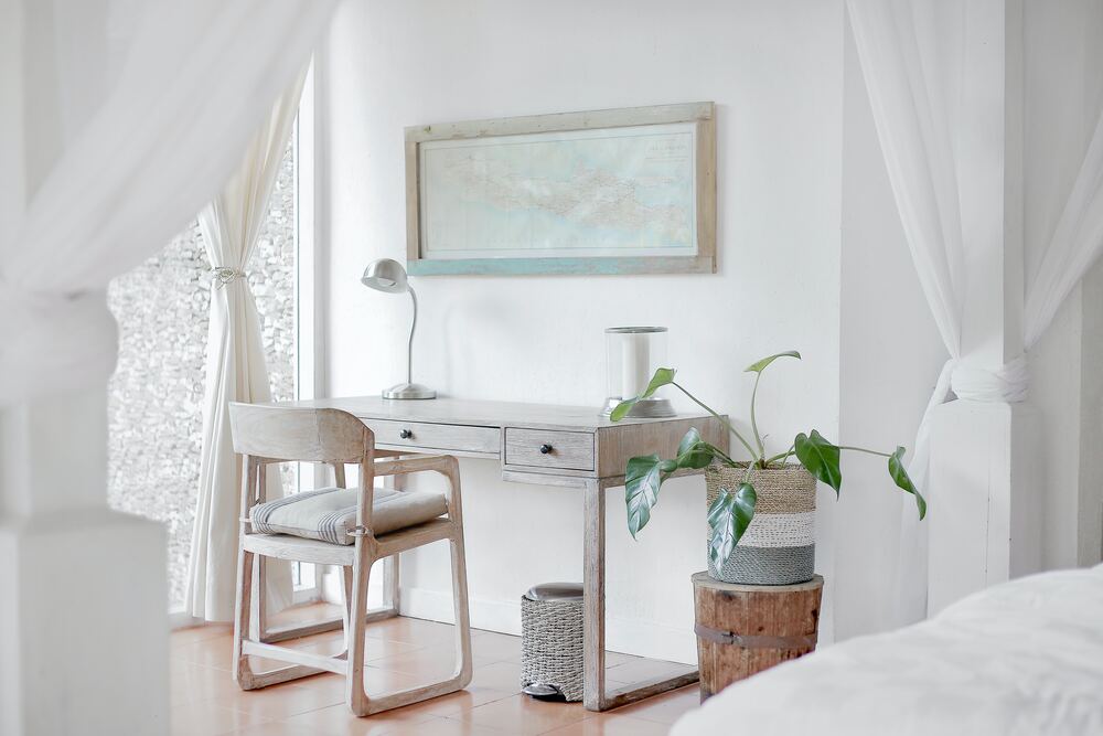 A minimalist aesthetic bedroom that has white curtains, a white bed, and neutral colored furniture made from natural materials. A potted plant sits beside the desk.
