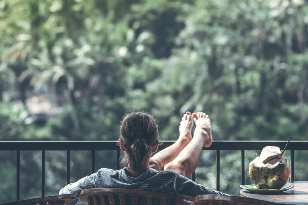 A barefoot woman kicking back while on vacation. She is relaxing outside with her legs up sitting next to a coconut beverage. This image represents the life you can live when you generate passive income. 