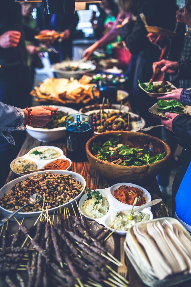 A close up image of a table with tons of food on it and people grabbing food. This is an image of a potluck.
